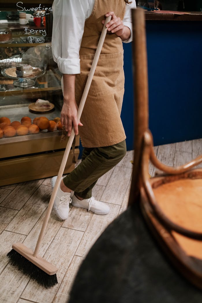 services-03 A person in an apron sweeps the wooden floor of a bakery with a broom, near pastries.