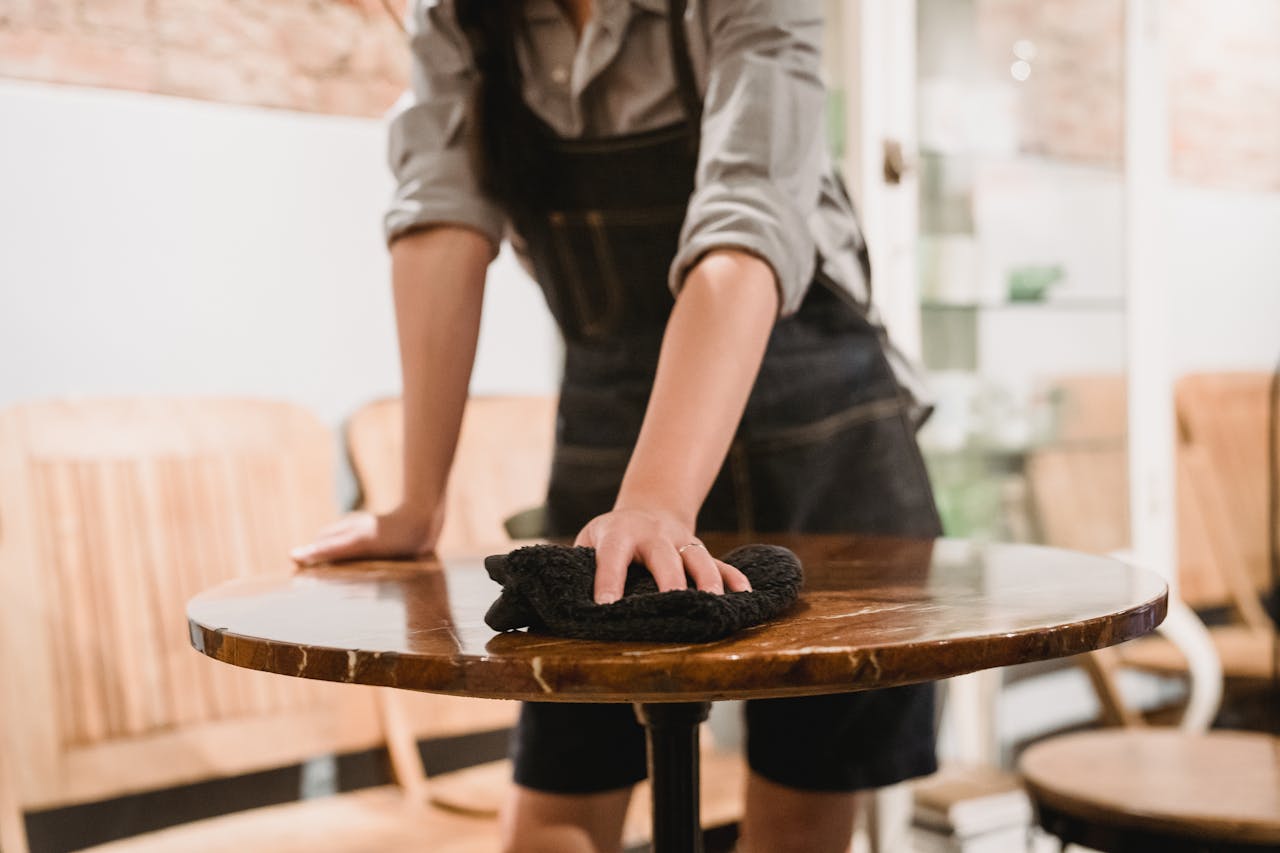 services-04 Person in apron cleaning a wooden table in a cozy café environment, focusing on hygiene.